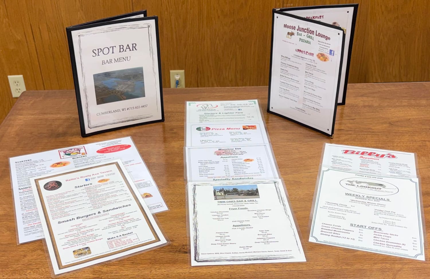 Finished menus designed for Manion Foods customers, standing and laid out on a table: Spot Bar of Cumberland, Moose Junction Lounge, Manitou Inn, Rollie&rsquo;s Rusty Axe Tavern, Twin Oaks Bar & Grill, Billy&rsquo;s and The Lookout